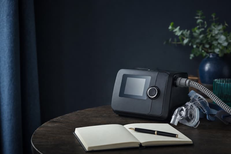Aparelho de CPAP sobre uma mesa escura, ao lado de um caderno aberto e de uma caneta