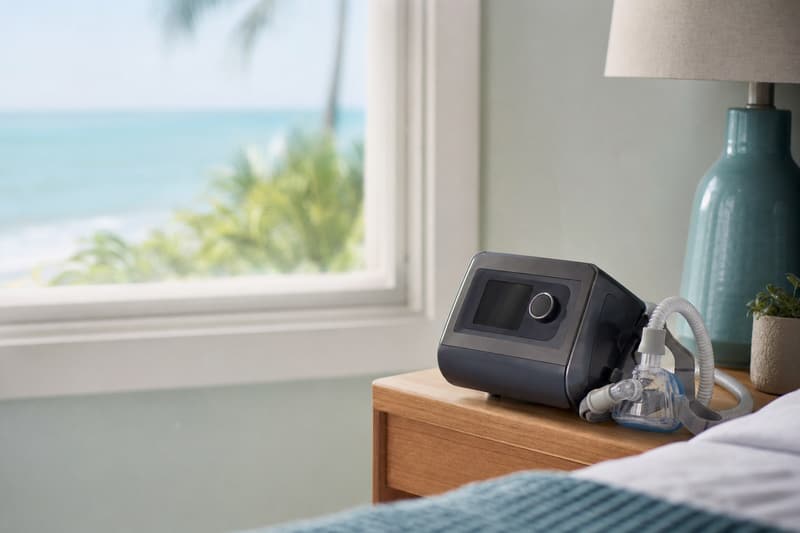 Aparelho de CPAP sobre um criado-mudo de madeira em um quarto claro, com o mar ao fundo pela janela