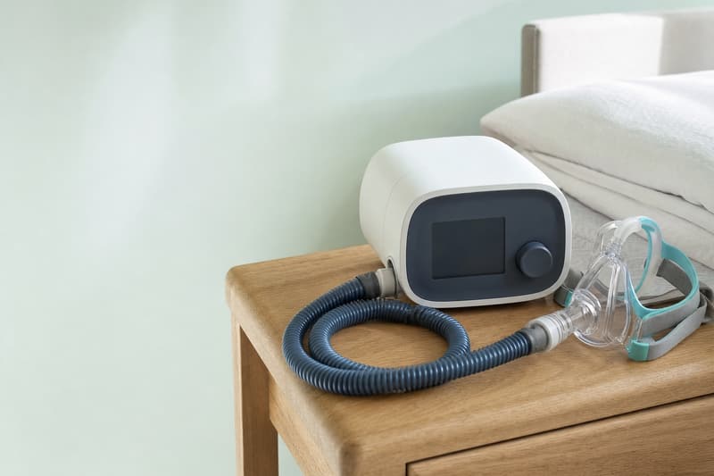 Aparelho de CPAP branco com a mangueira e a máscara nasal sobre um criado-mudo de madeira clara